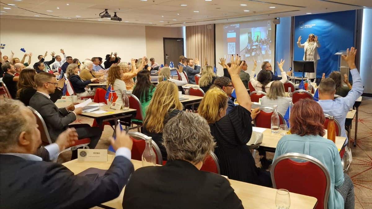 A group of people are seated in a conference room, raising their hands. A person stands at the front near a large screen, addressing the audience.