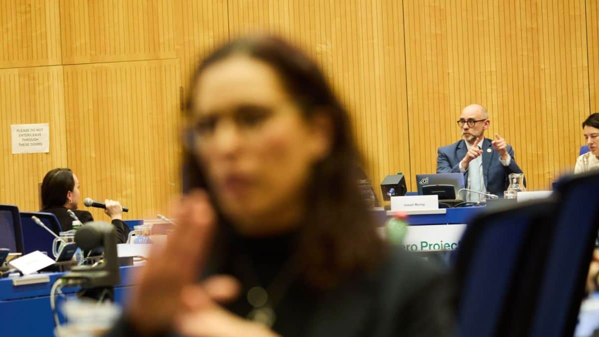 People are seated at conference tables in a room with wooden walls. A person in the foreground is speaking, while others listen attentively. A sign reads "Please do not use cameras.