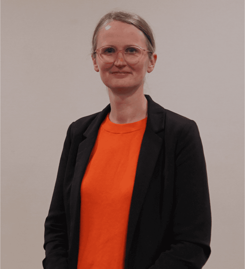 Person wearing glasses, an orange top, and a black blazer, standing in front of a neutral background.