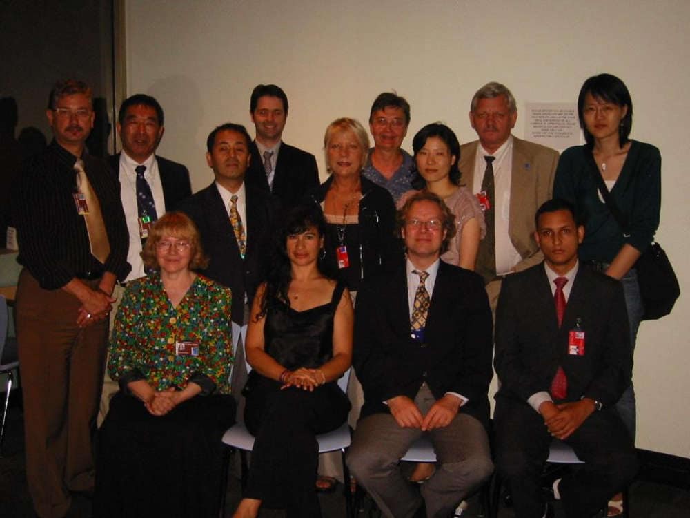 A group of thirteen people in formal attire pose together indoors against a plain wall.