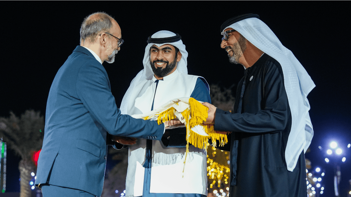 Three men in a formal outdoor setting, two wearing traditional Middle Eastern attire presenting an object to a man in a suit.