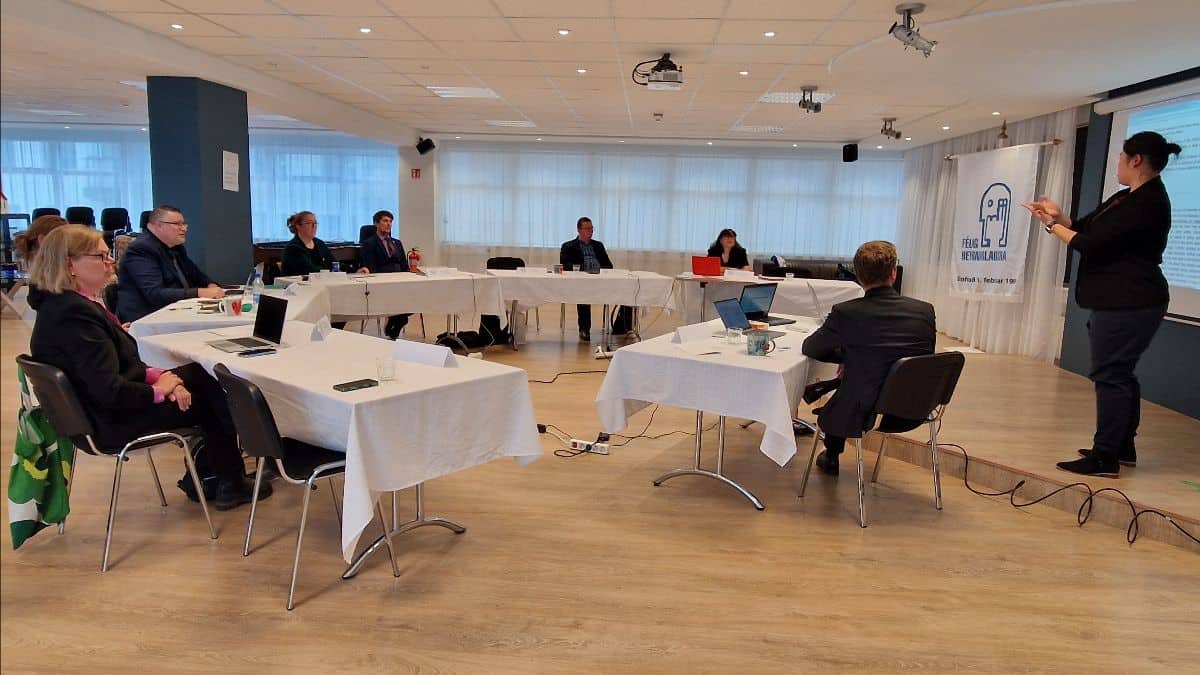 A business meeting with seven people sitting around tables in a conference room setting. One person is standing and presenting, using a screen.