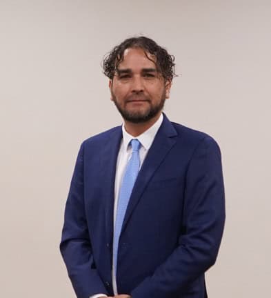 A man in a blue suit and tie stands against a plain background.