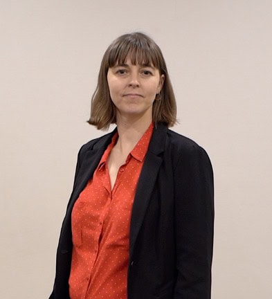 Person with shoulder-length brown hair wearing a red shirt and black jacket, standing against a plain background.