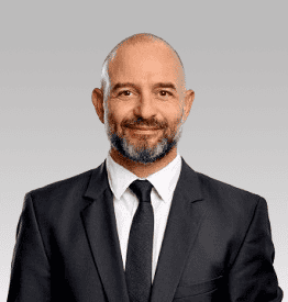 A bald man with a beard, wearing a dark suit and tie, stands against a plain gray background, smiling.