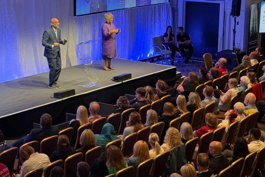 A man speaks on stage next to a woman interpreter, facing an audience seated in rows at an indoor event.