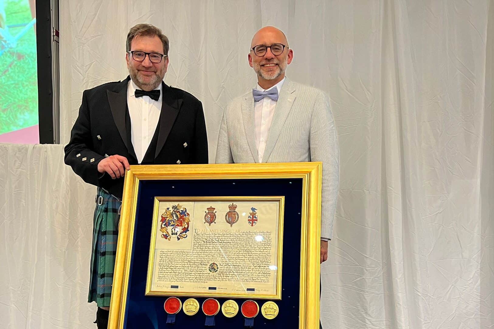 Two men stand on a stage with a framed certificate featuring gold seals, resting on a chair in front of them. One man wears a kilt; the other is in a light suit and bow tie.
