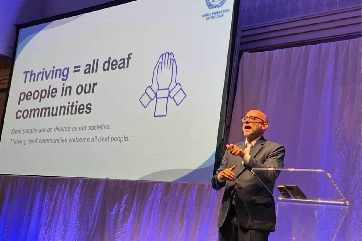 A man in a suit uses sign language on stage beside a screen displaying a slide about supporting deaf communities, with the World Federation of the Deaf logo at the top.