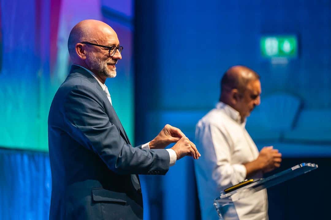 Two men stand at a podium; one in a suit speaking and gesturing, the other in a white shirt looking down, with a blue-lit background behind them.