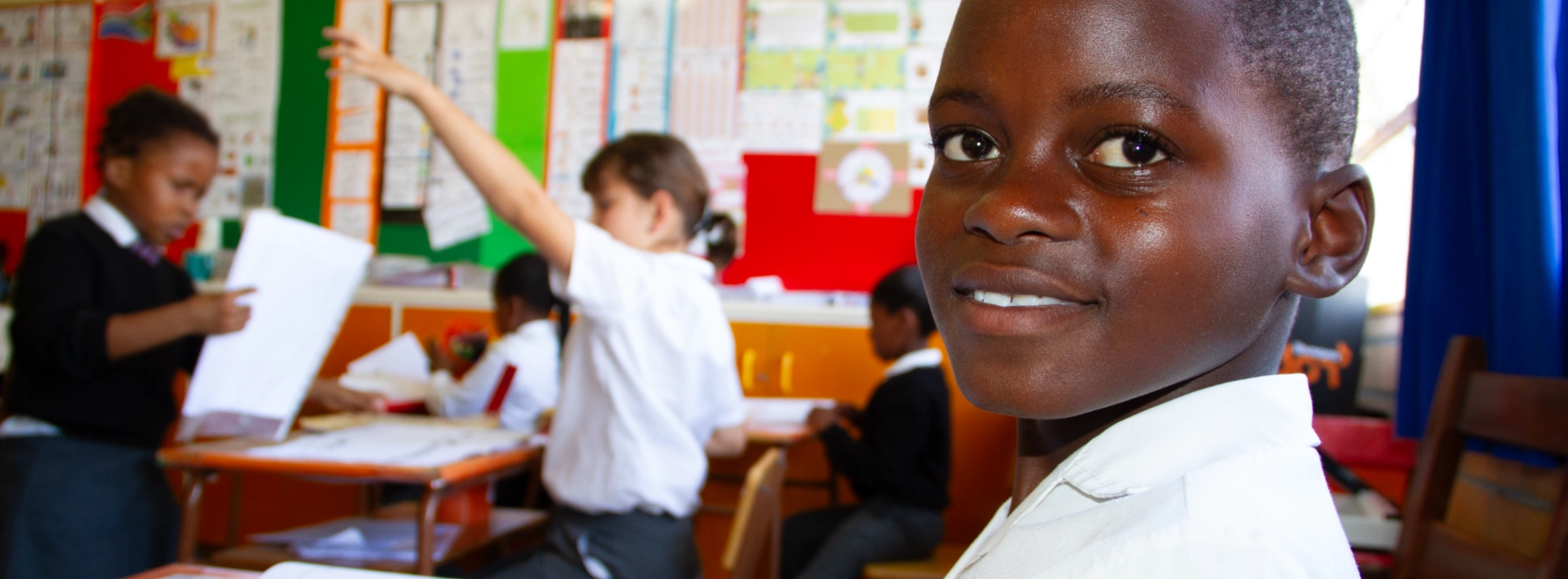 A student smiles in a colorful classroom while other children engage in activities at their desks.