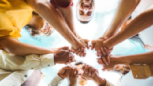 A group of people stand in a circle, reaching their hands toward the center, joining them together against a blue sky background.