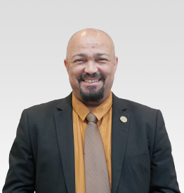 A man with a bald head and goatee, wearing a black suit, brown tie, and yellow shirt, stands against a plain light background, smiling at the camera.