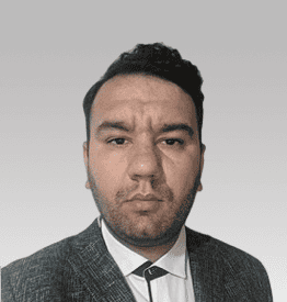 A man with short dark hair, wearing a grey suit jacket and white shirt, looks directly at the camera against a plain light grey background.