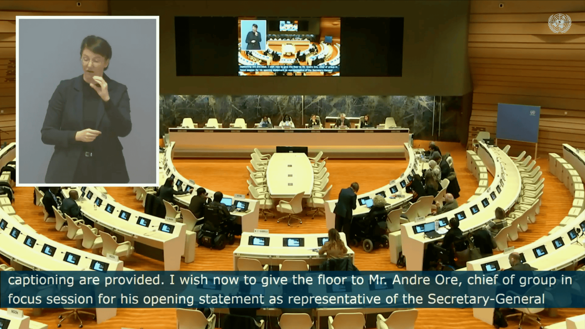A conference room with a large oval table and delegates seated. A screen displays a speaker. A sign language interpreter is shown in an inset box.