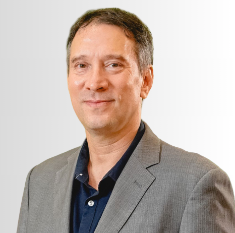 Man in a gray suit and dark shirt smiling at the camera against a plain background.
