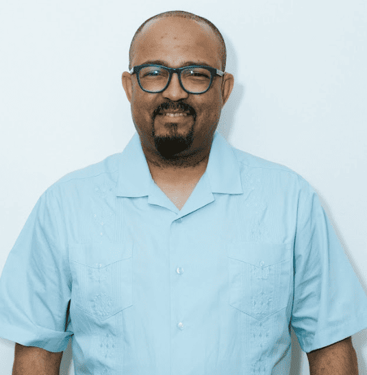 A man with glasses and a beard wearing a light blue shirt stands against a plain white background, smiling at the camera.