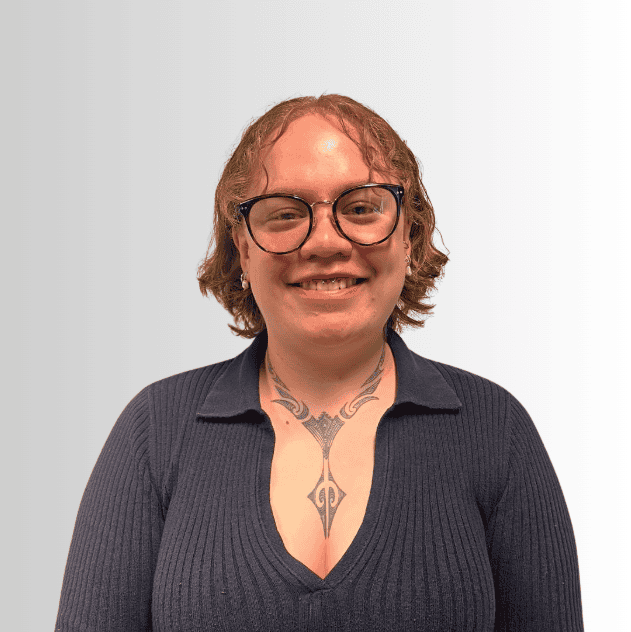 A person with short brown hair, glasses, and a visible chest tattoo is smiling, wearing a dark ribbed shirt, in front of a light gradient background.