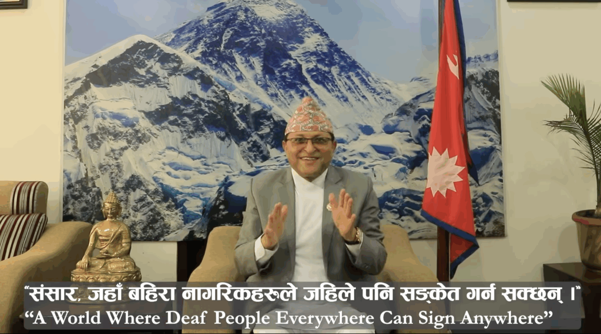 A man in traditional attire sits in front of a Nepal flag and mountain mural, gesturing with his hands. A Buddha statue is on the table. Caption: “A World Where Deaf People Everywhere Can Sign Anywhere.”.