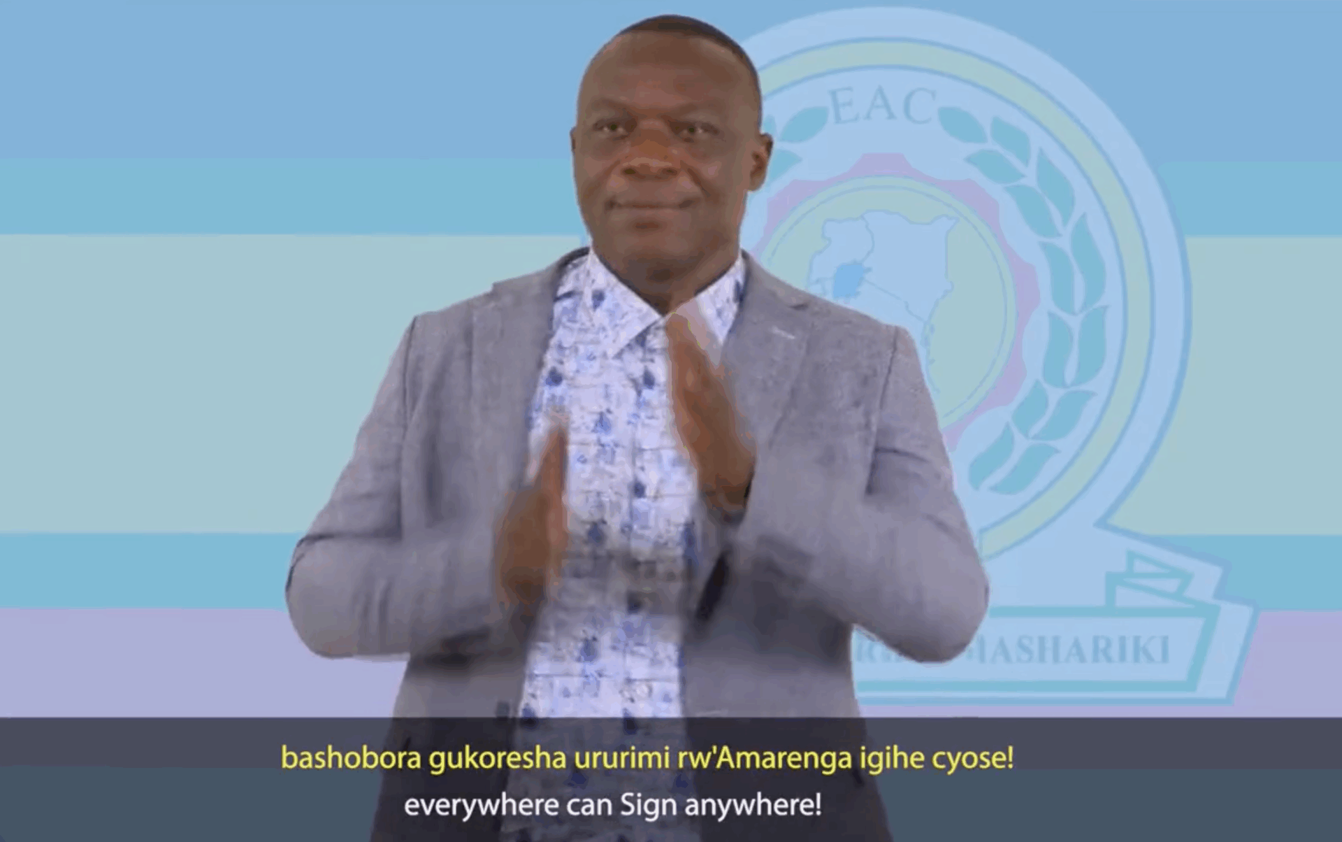 A man in a gray blazer and patterned shirt uses sign language, with an East African Community logo in the background and subtitles promoting sign language use.