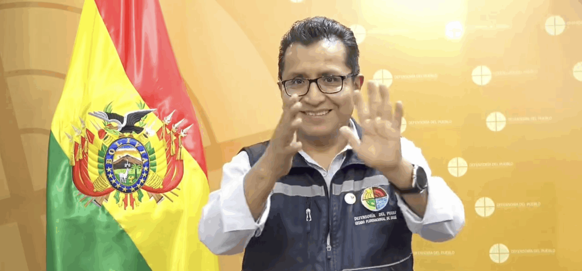 A man stands in front of a Bolivian flag, wearing a vest with official badges, using sign language with a smile.
