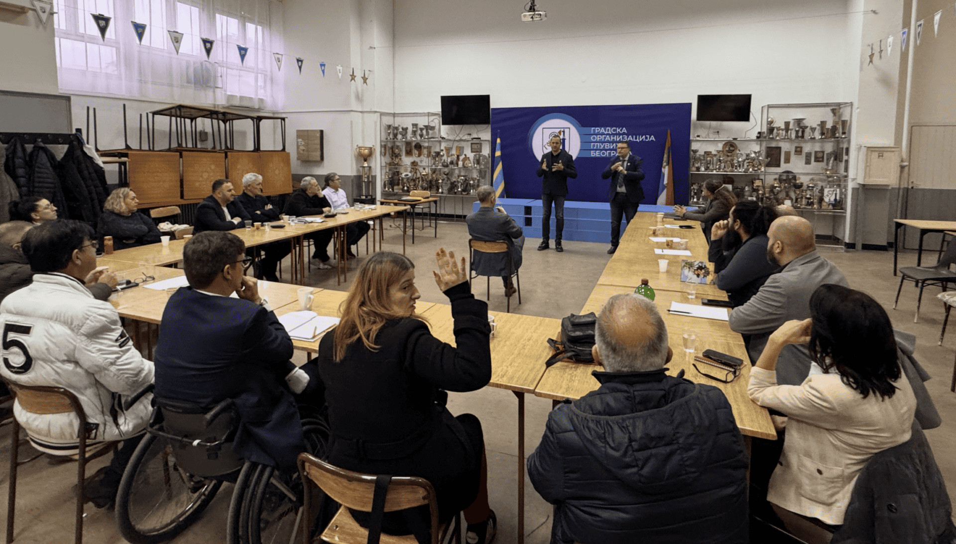A group of people sit round tables in a meeting room, listening to two standing speakers in front of a blue backdrop with text.