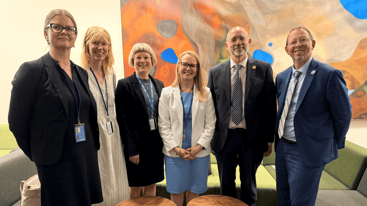 Six professionals in formal attire stand together indoors in front of a colourful abstract wall art, posing for a group photo.