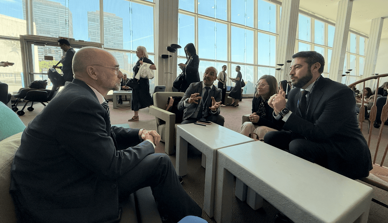 Four people in business attire are seated and engaged in discussion in a bright, modern lobby with large windows and city buildings visible outside.