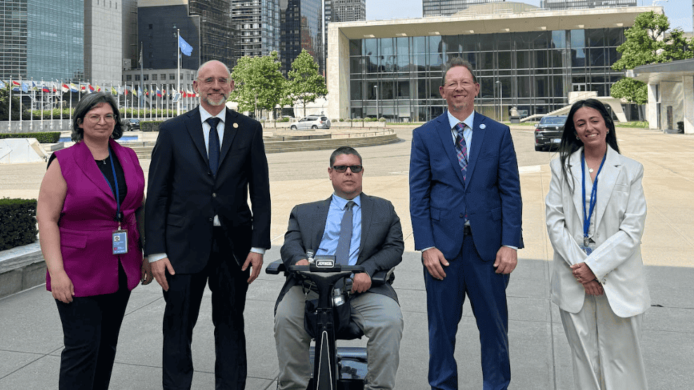 Five people, including one person in a mobility scooter, pose for a group photo outside the United Nations Headquarters, with city buildings and flags visible in the background.