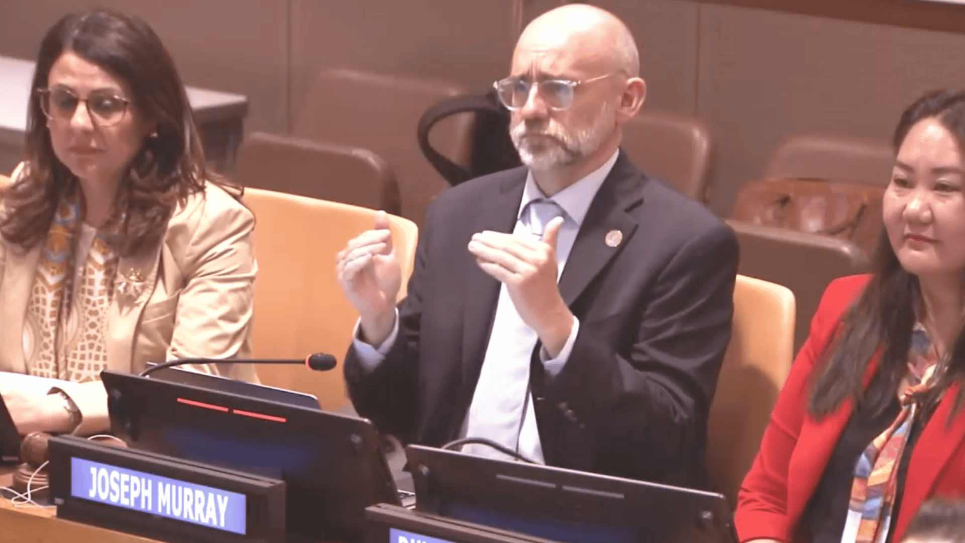 Three panellists sit at a conference table with nameplates; the man in the centre gestures with hands raised, while the other two face forward.