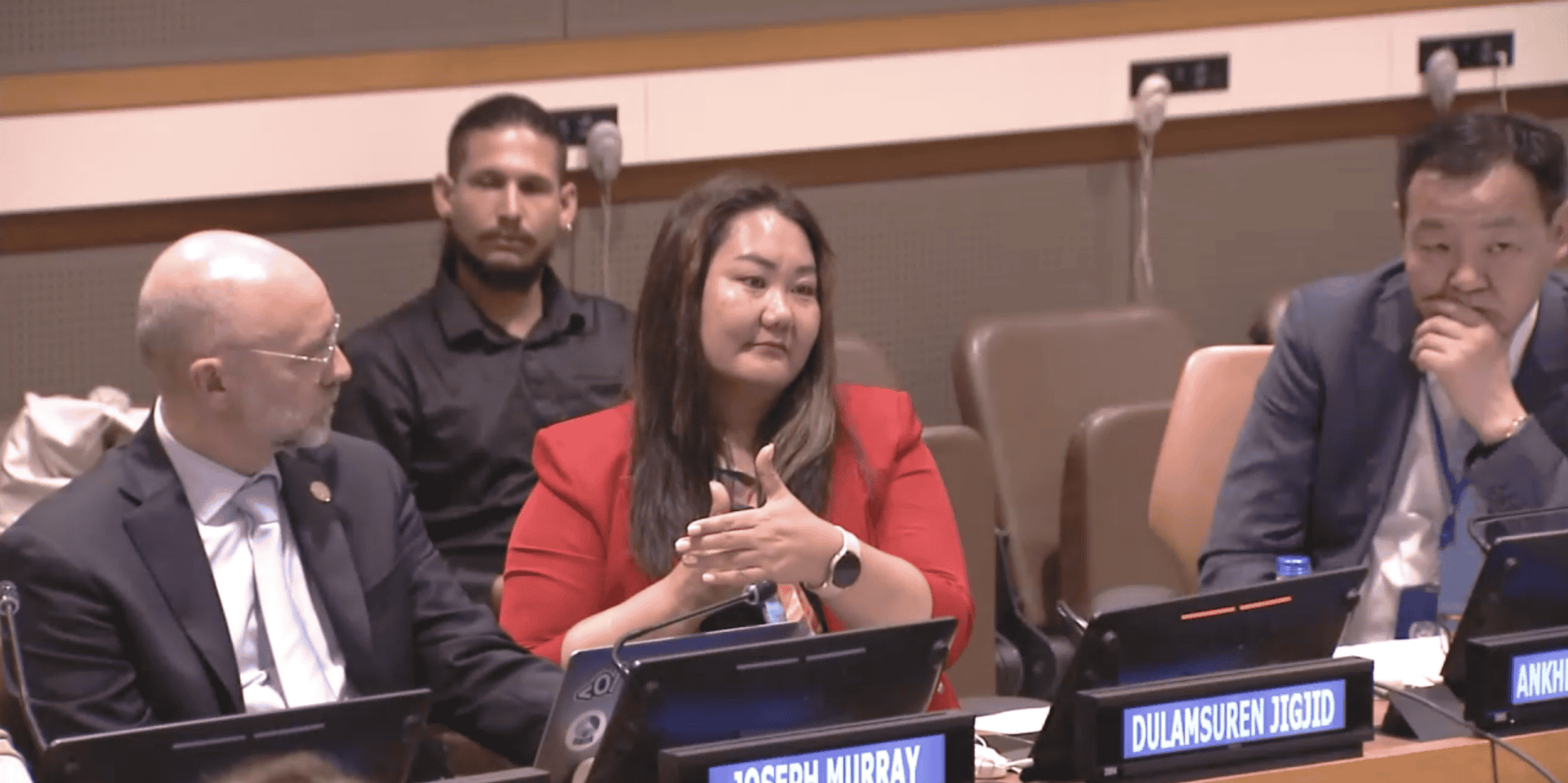 Four people sit at a conference table, some with laptops in front of them. One person in a red jacket is speaking, whilst others listen. Name placards and microphones are visible.