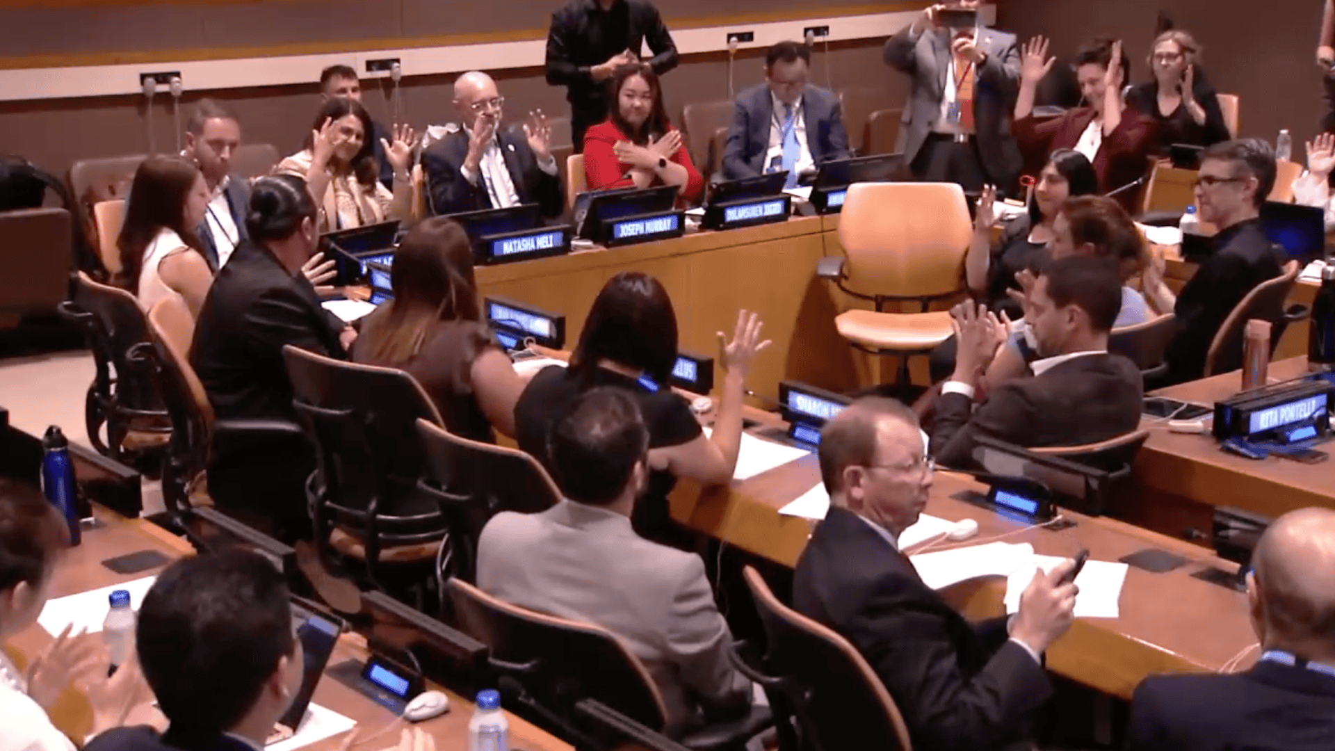 A group of people seated around a conference table raise their hands during a formal meeting in a large room.
