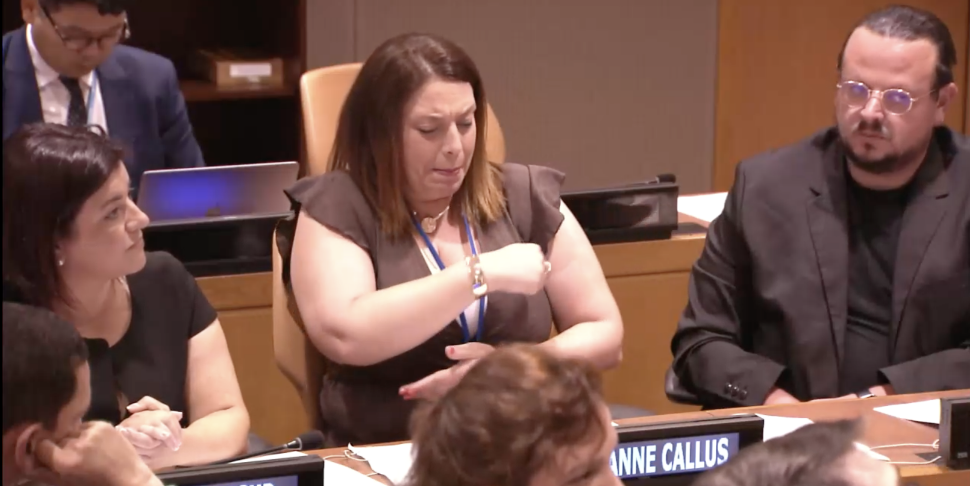 A woman uses sign language while seated at a conference table, with two other people beside her and nameplates visible in front.