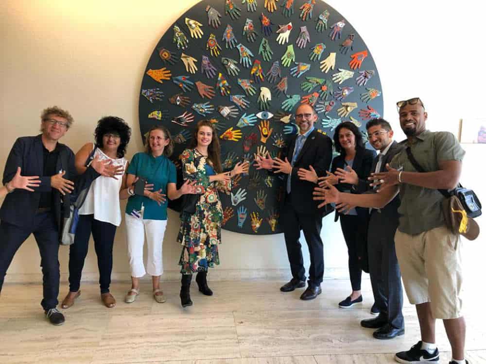 A group of people stand smiling in front of a large circular artwork featuring colorful handprints.