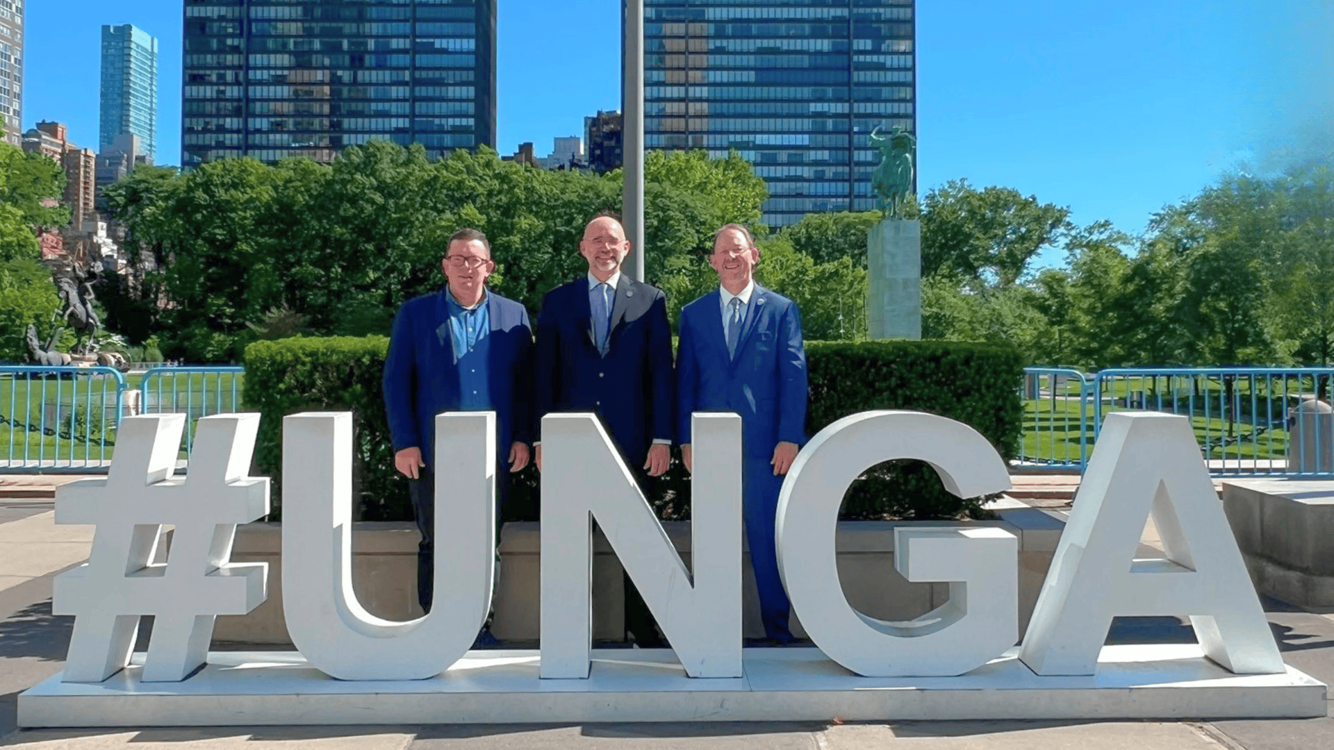 Three men in suits stand behind large white "#UNGA" letters outdoors with buildings and trees in the background.