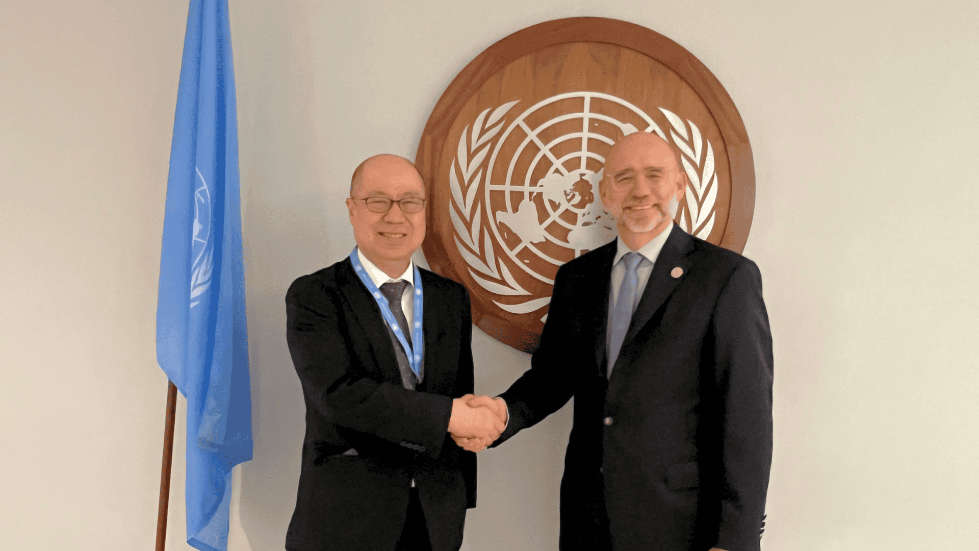 Two men in suits shake hands in front of a United Nations emblem and flag.