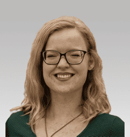 A person with wavy hair and glasses smiles in front of a neutral background.
