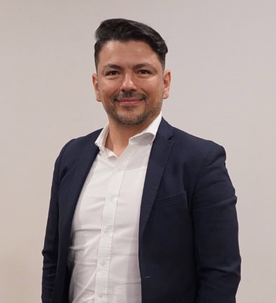 A person in a suit and white shirt stands against a plain background, facing the camera, with a slight smile.