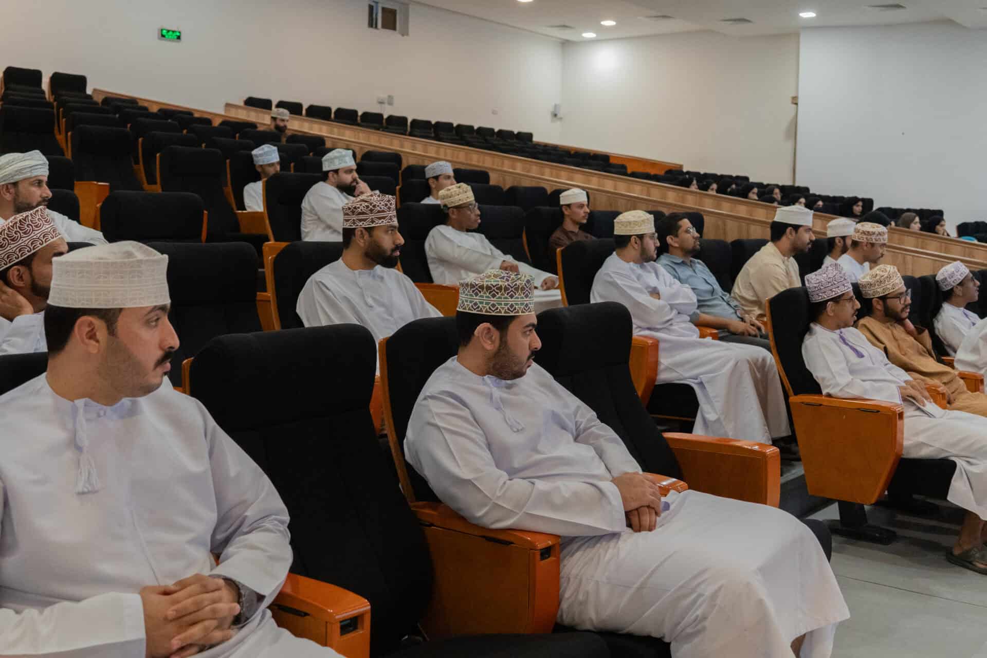 Men in traditional Omani attire sit in black and orange seats in a lecture theatre, attentively facing forward during a presentation or event.