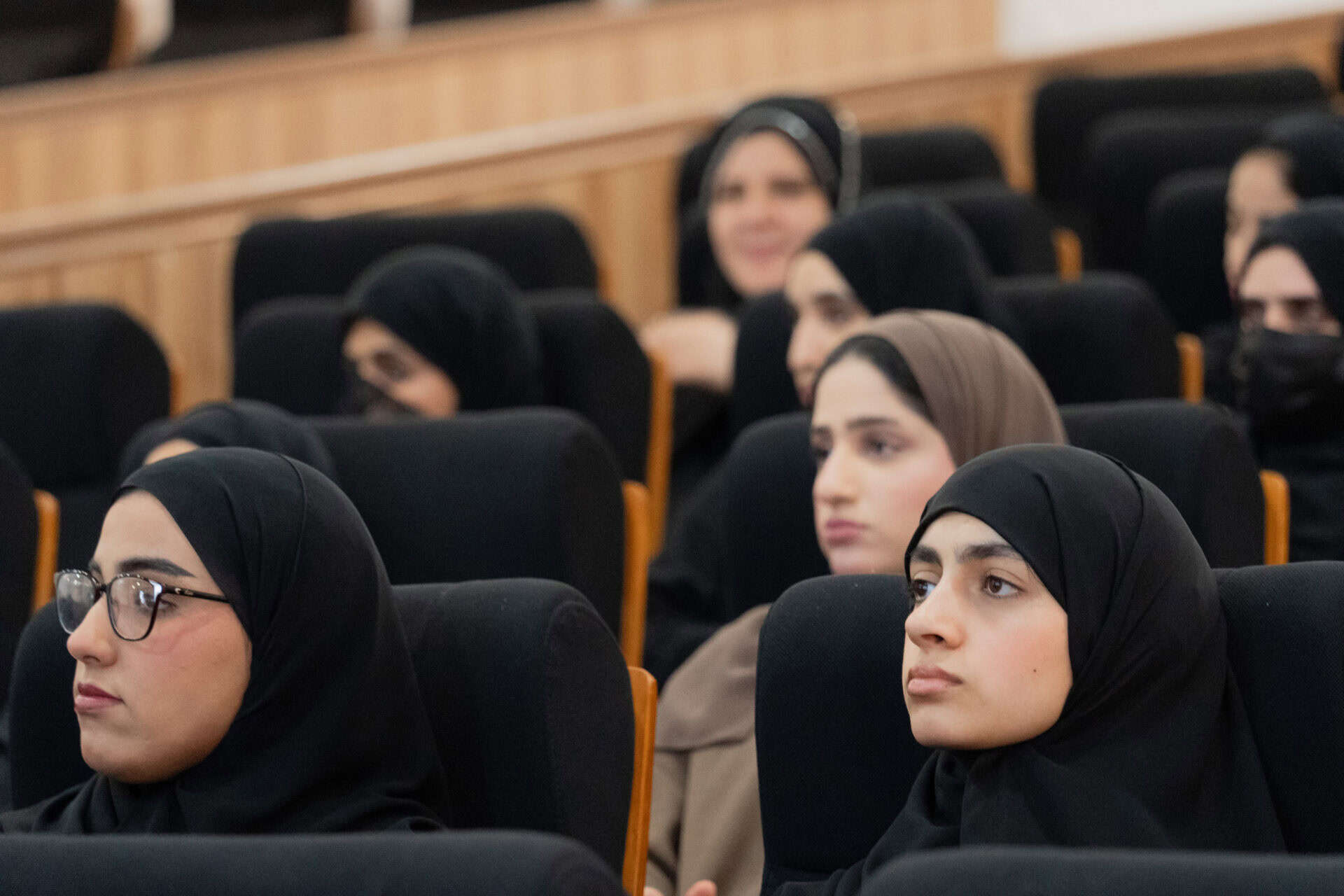 Several women wearing hijabs are seated in an auditorium, attentively listening.