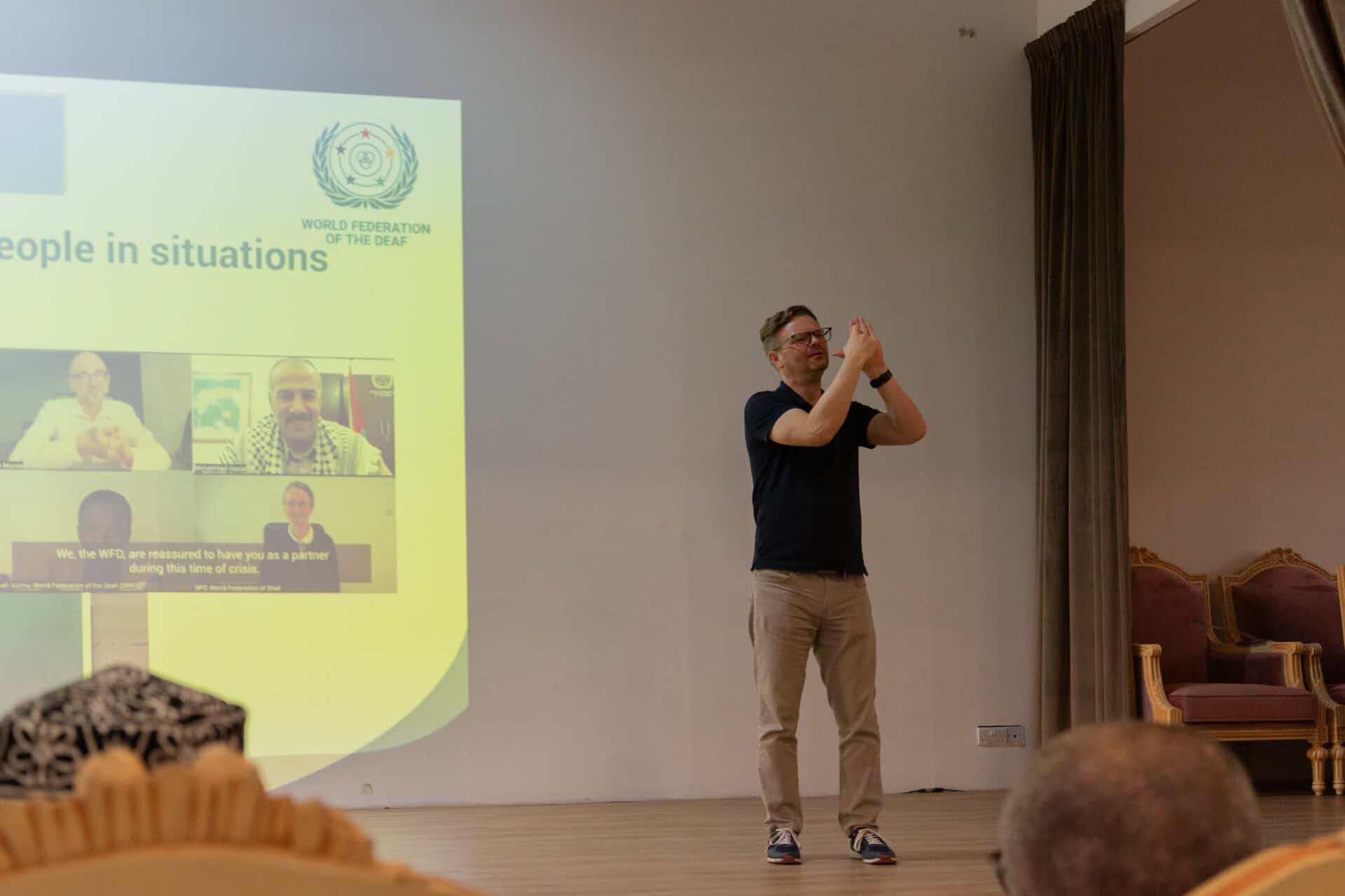A man stands on stage signing in front of a projected presentation slide, with seated audience members in the foreground.