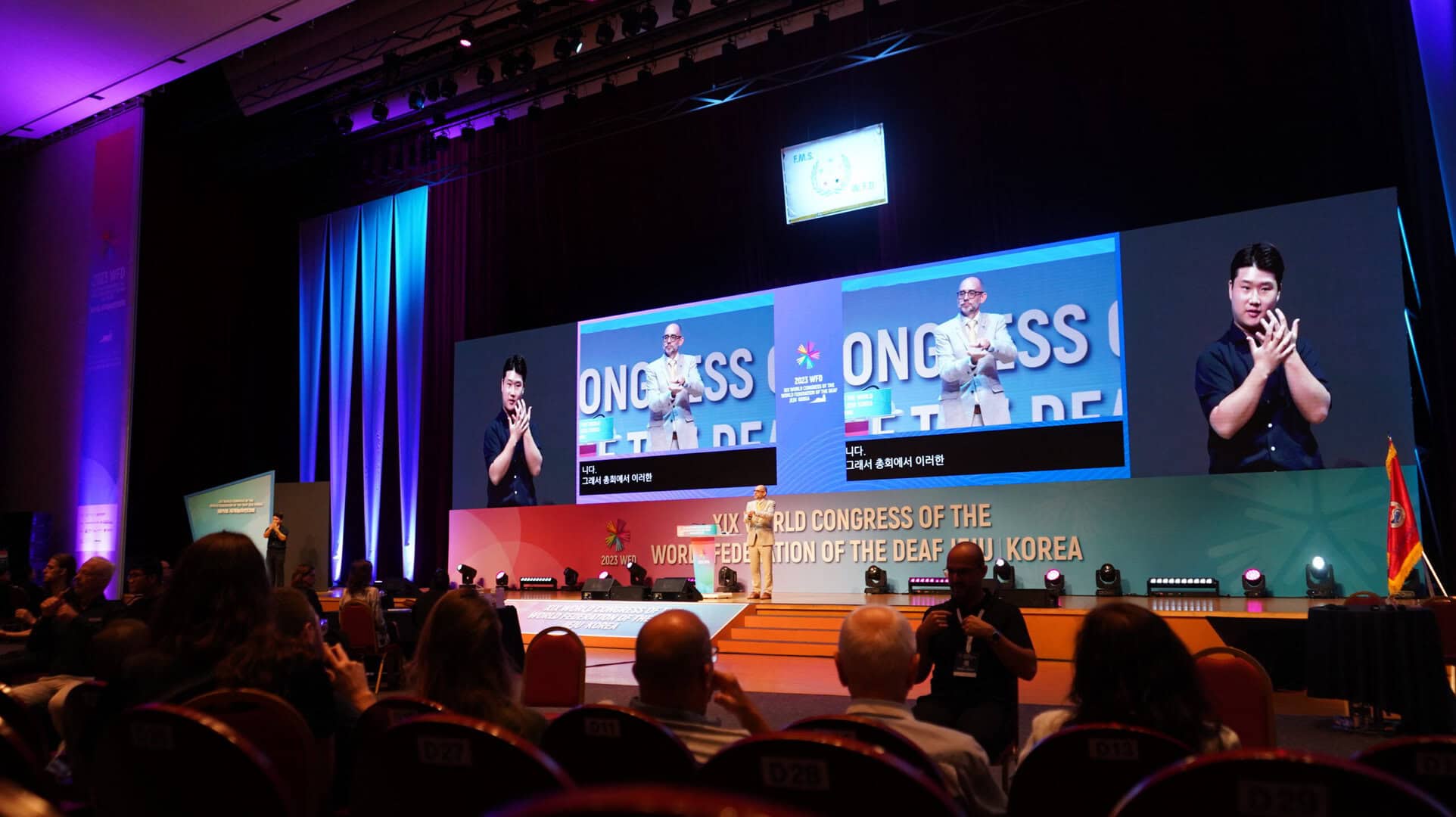 Large conference with a speaker using sign language on stage. Two screens display the speaker, and audience members are seated in rows.
