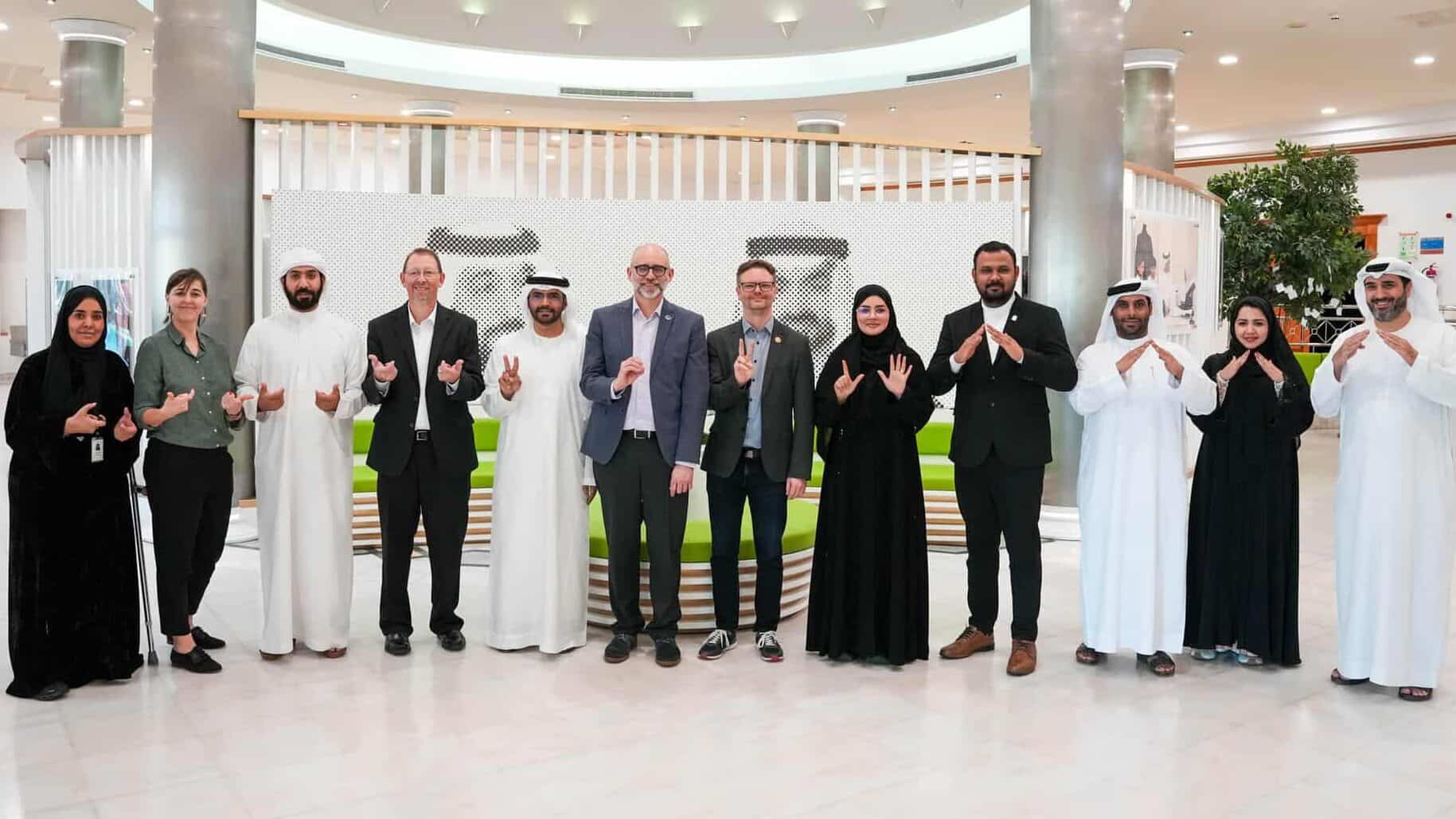 A group of twelve people, men and women in business attire and traditional clothing, stand indoors in a semicircle, smiling and making hand signs.