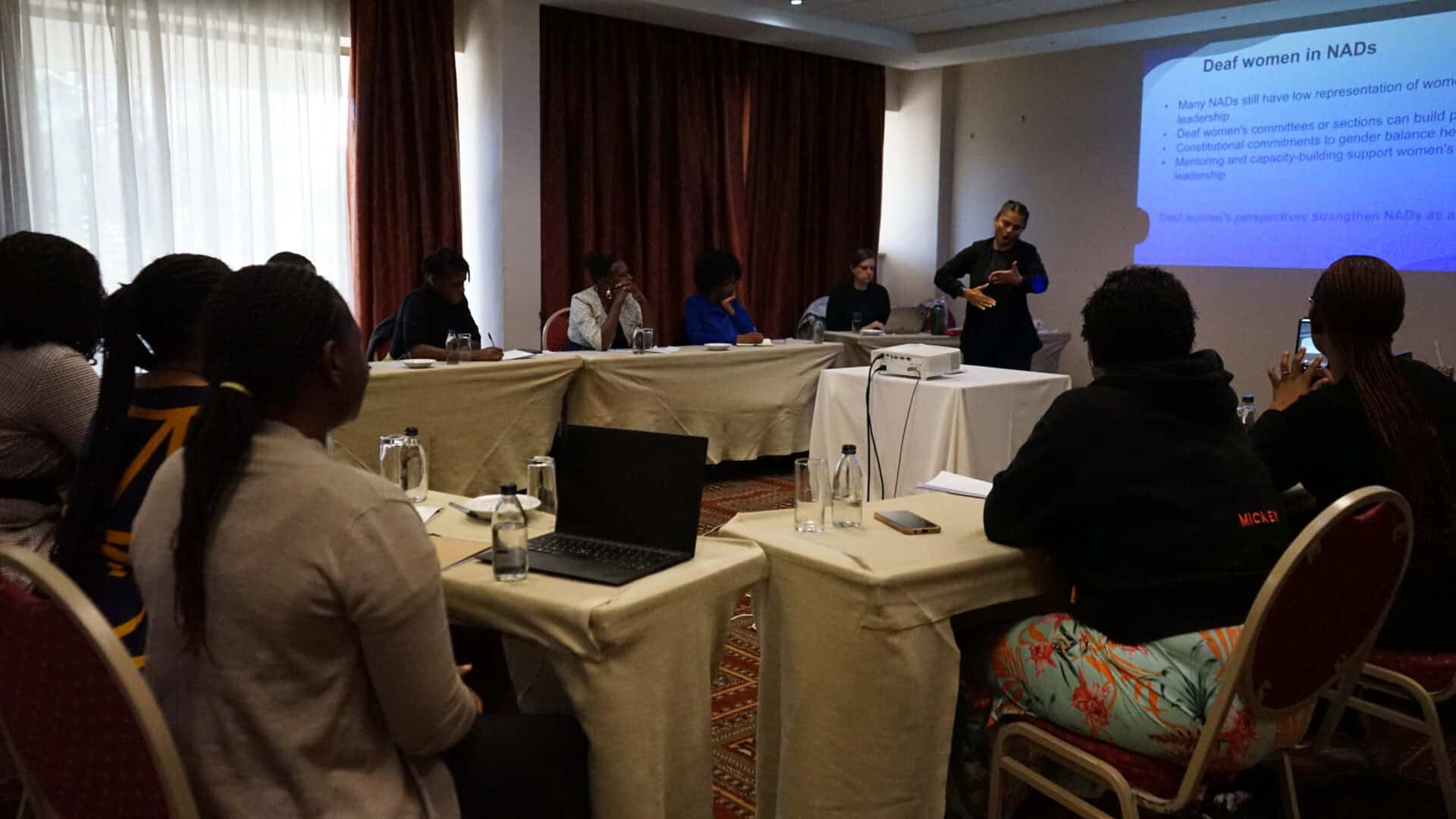 A group of people sit around tables in a conference room whilst a presenter stands at the front, gesturing towards a projected presentation on a screen.