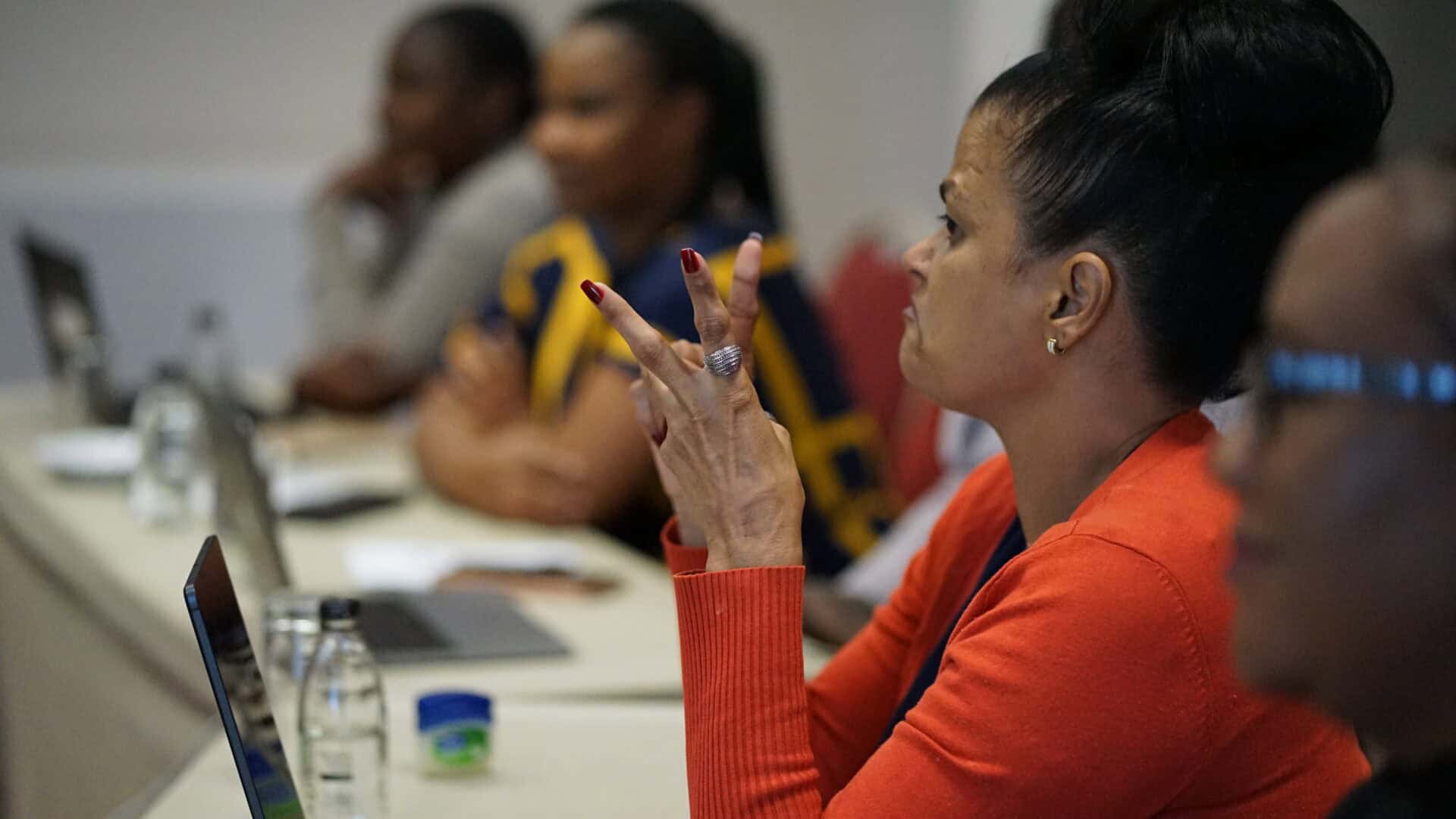 A woman in an orange jumper sits at a table with a laptop, gesturing with her hands, while others listen in a blurred background at a meeting or workshop.