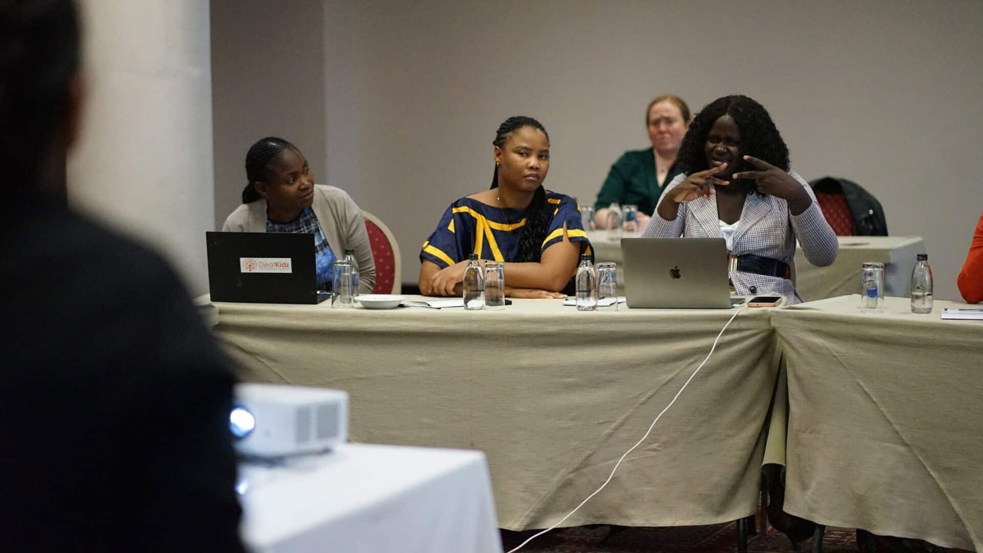 Four women sit at a conference table with laptops and water bottles; one woman is gesturing while speaking, and others listen attentively.