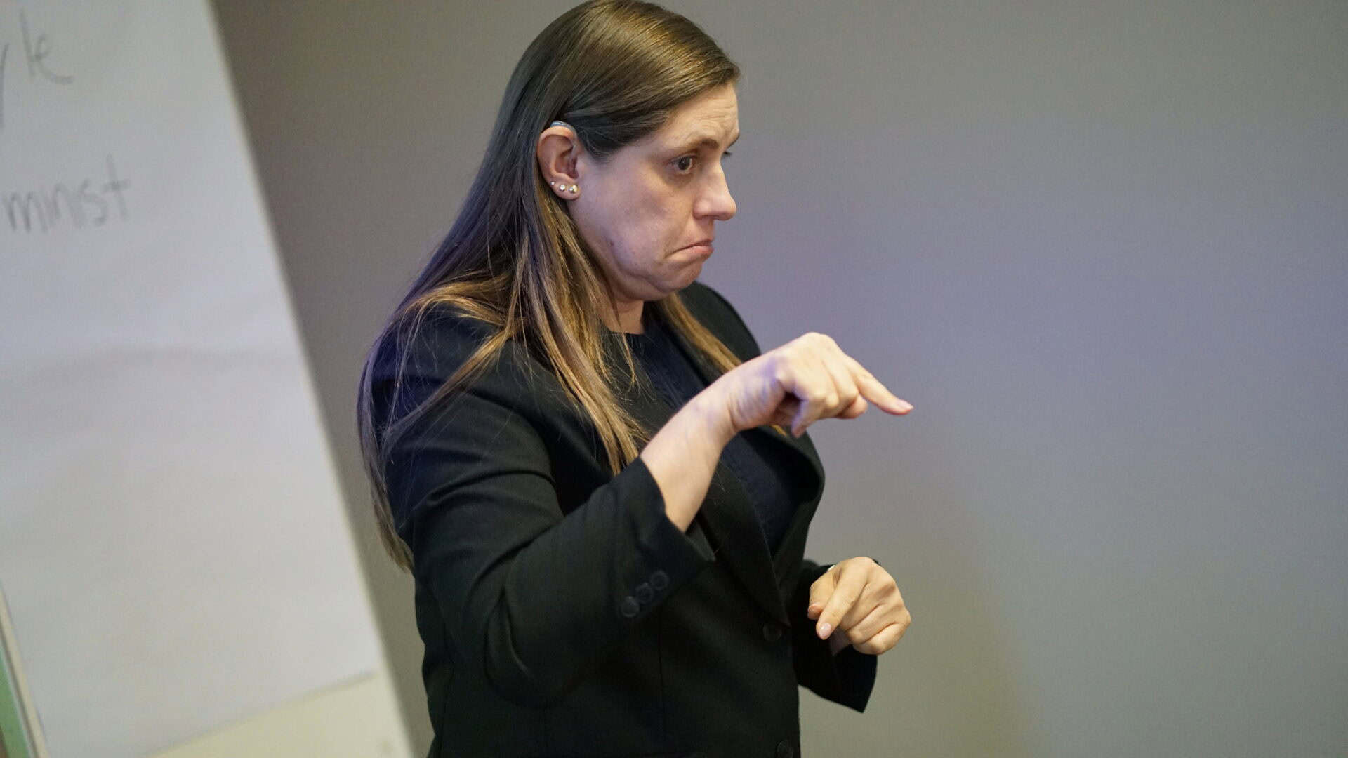 A woman with long brown hair in a black blazer uses sign language in front of a flipchart.