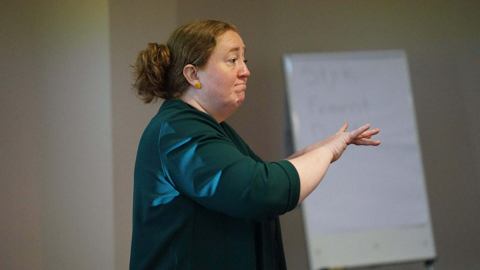 A woman with brown hair tied back gestures with one hand whilst standing indoors near a blurred flip chart.
