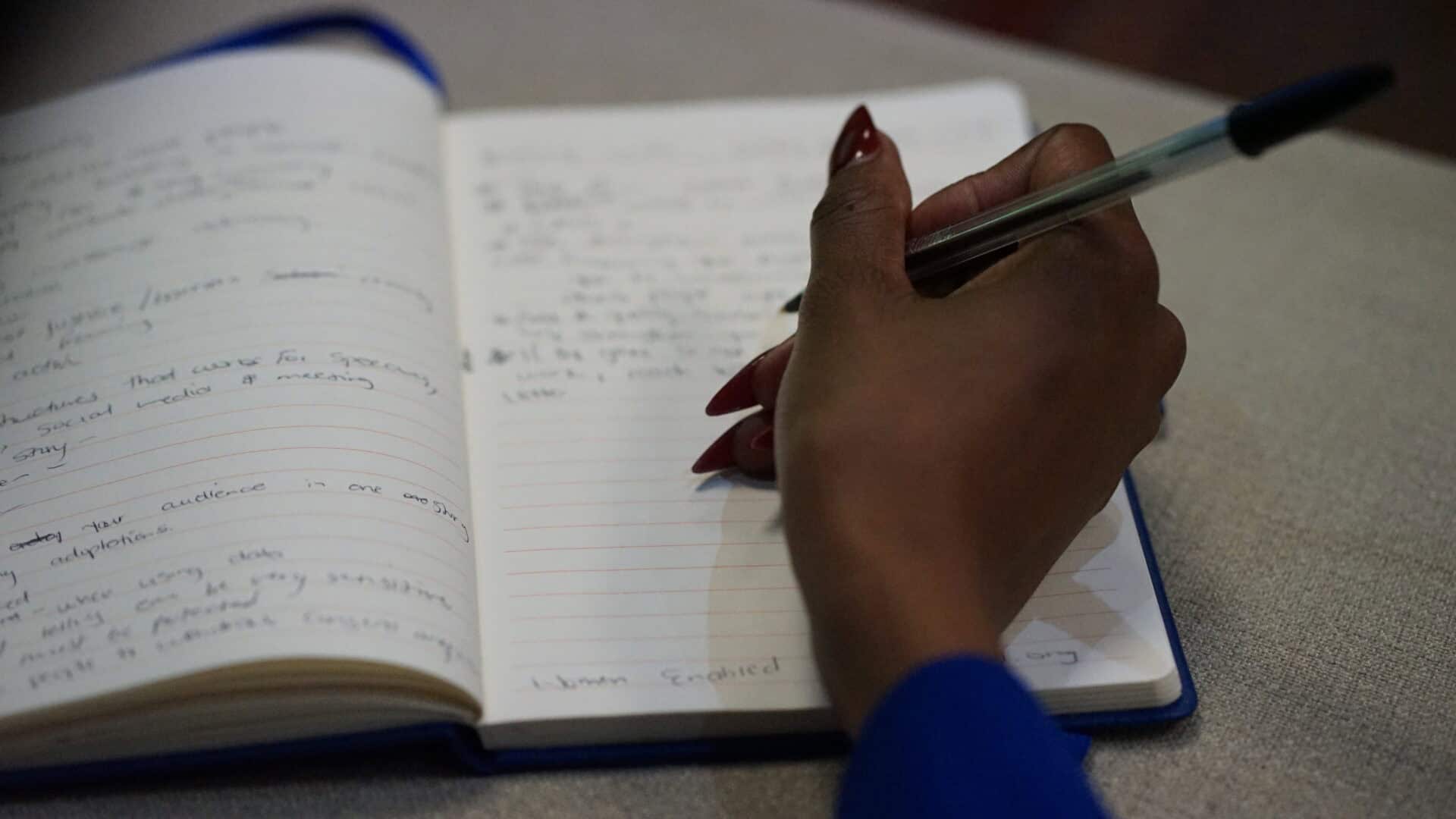 A person with red nails writes in a notebook with a blue pen; handwritten notes are visible on the open pages.
