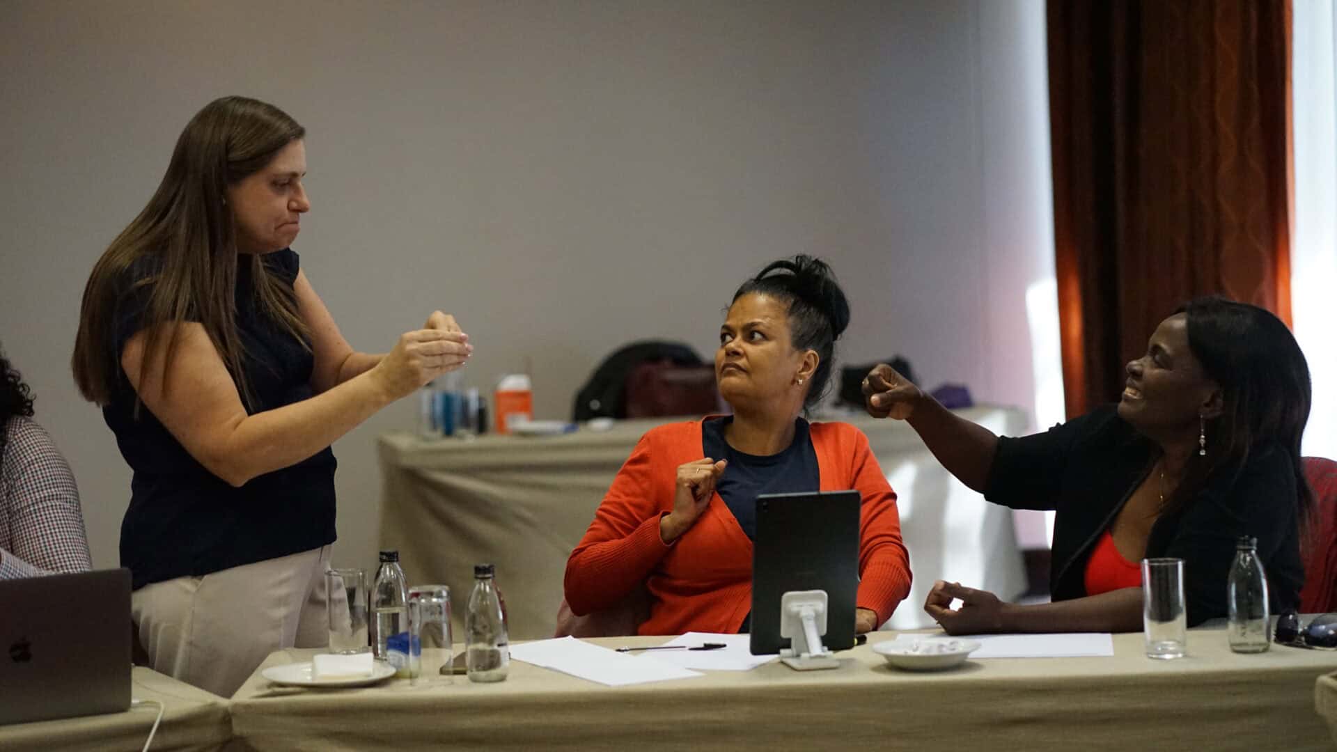 Three women sit at a table in a meeting room, engaging in animated conversation; one woman stands, gesturing with her hands.