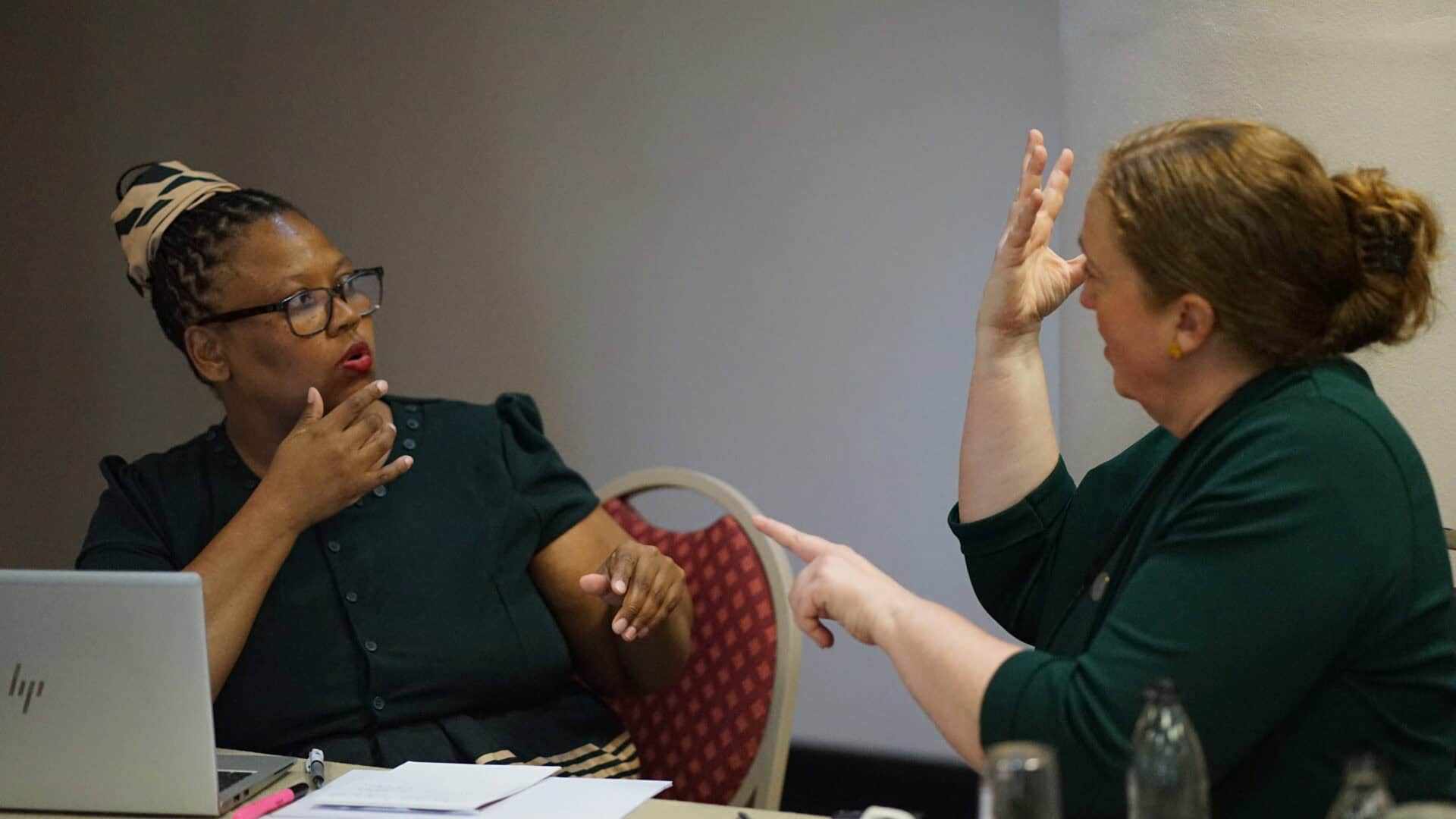 Two women sit at a table in discussion; one gestures with her hand raised whilst the other touches her chin, with a laptop and papers in front of them.